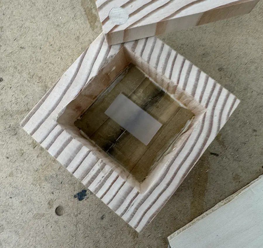 Small wooden box with two small darker-colored planks on the bottom of its cavity and a small piece of acrylic on top of them.