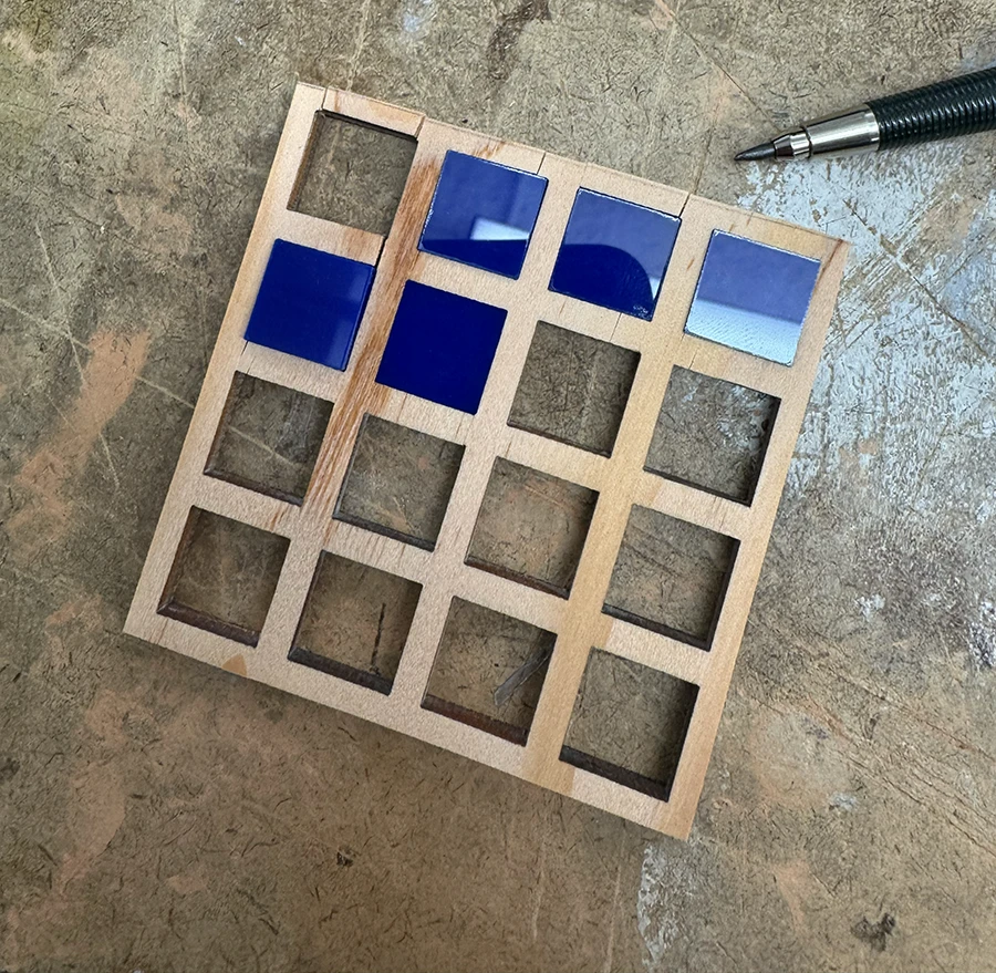Small wooden square with a 4 by 4 grid of squares, filled with acrylic pieces. Cracks on the perimeter of the wood are visible.