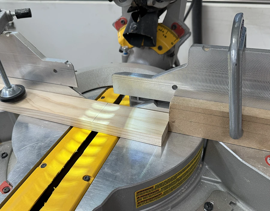 A compound miter saw with a block clamped to its side and another ready to be cut.