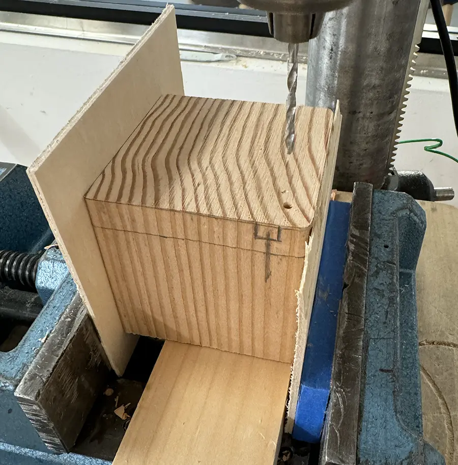 A cube-like piece of wood being drilled on the bench press, on its top right corner.