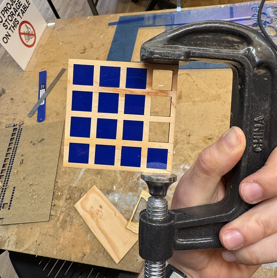 Small wooden square with a 4 by 4 grid of squares, filled with acrylic pieces. Being held with a clamp.