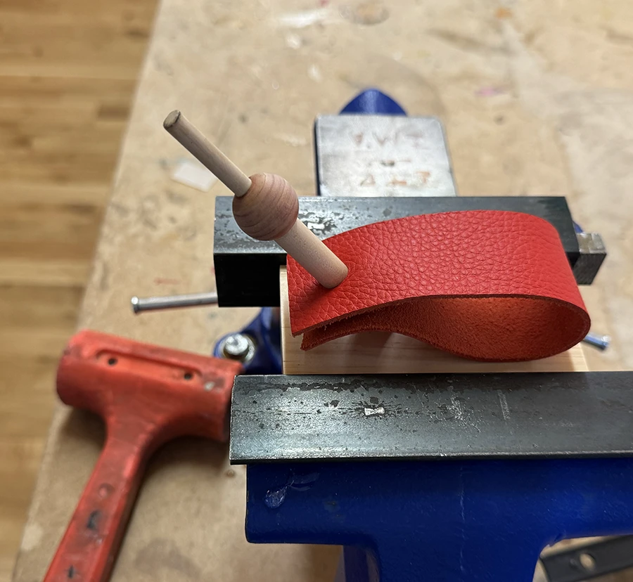 A block of wood with a diagonal dowel coming out of it, clamped down on a vise.