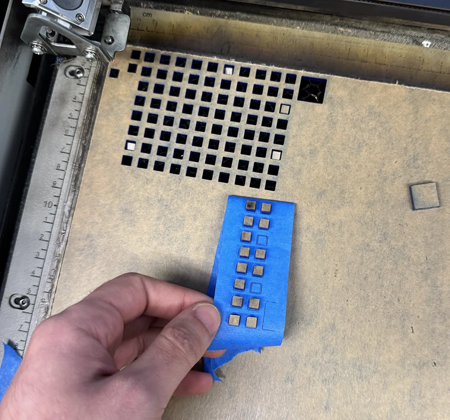 Acrylic on the bed of a laser cutter. A strip of tape is being used to take out small square cutouts.