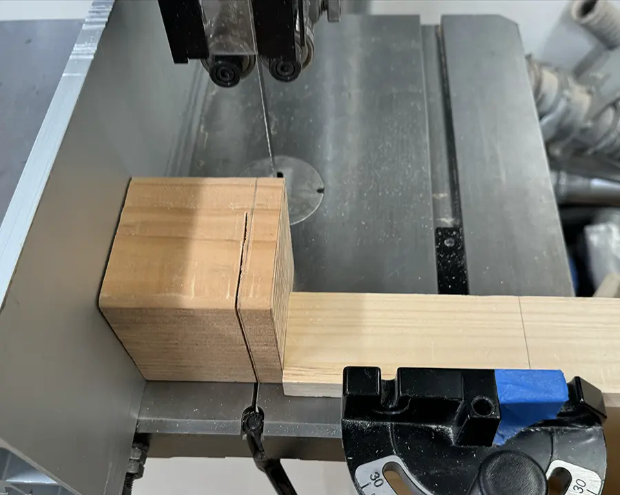 A cube-like piece of wood being cut on the band saw. Another adjacent piece of wood holds the piece being cut steady.