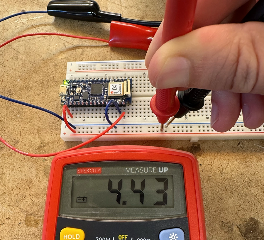 Arduino Nano IoT 33 on the edge of a breadboard. Hands use a multimeter to measure two points on the breadboard, connected to vcc and ground. It reads 4.43V