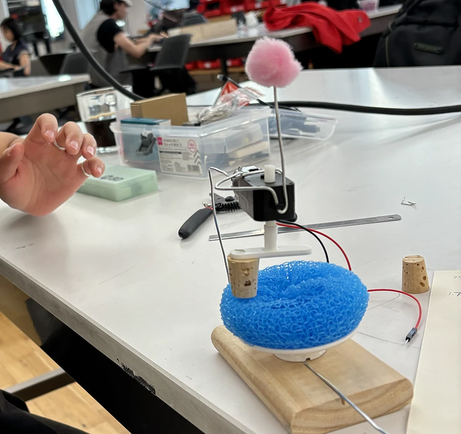 Small structure with a wooden base, a plastic dish spong facing upwards, an a motor held right over it with a metal wire. A small plastic t-shaped object mounts a cork to the motor spindle, and hangs over the sponge.