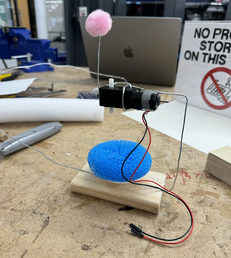 Small structure with a wooden base, a plastic dish spong facing upwards, an a motor held right over it with a metal wire.