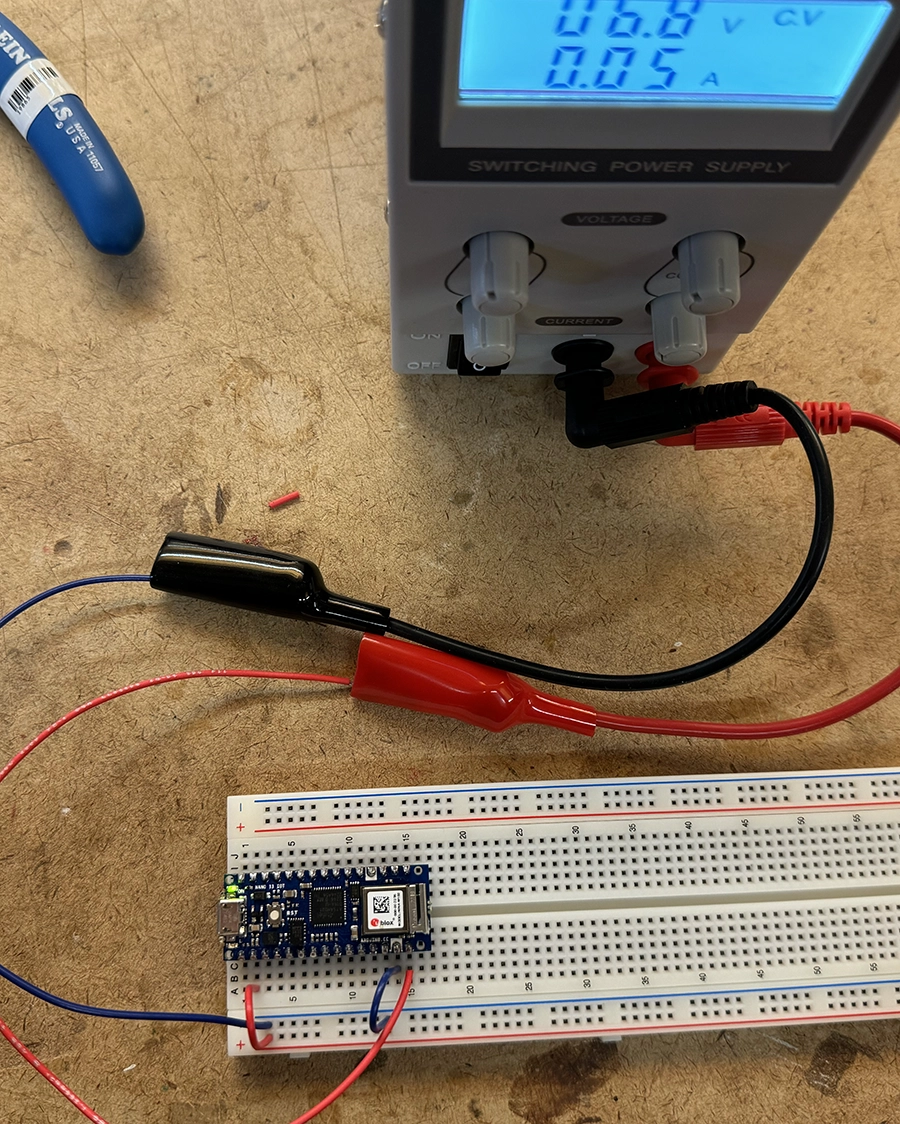 Arduino Nano IoT 33 on the edge of a breadboard. Connected with red and blue cables to a variable power supply that reads 6.8V.