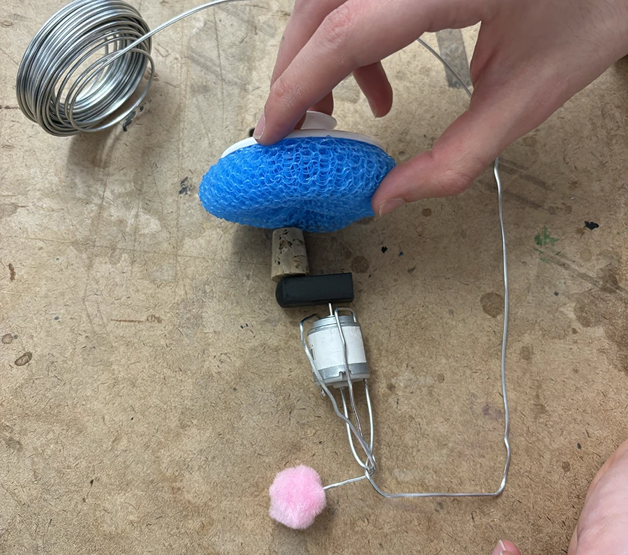 A small dc with an eraser stuck to its axis, and a small cork touching the eraser on one side and a blue plastic dish sponge on the other..